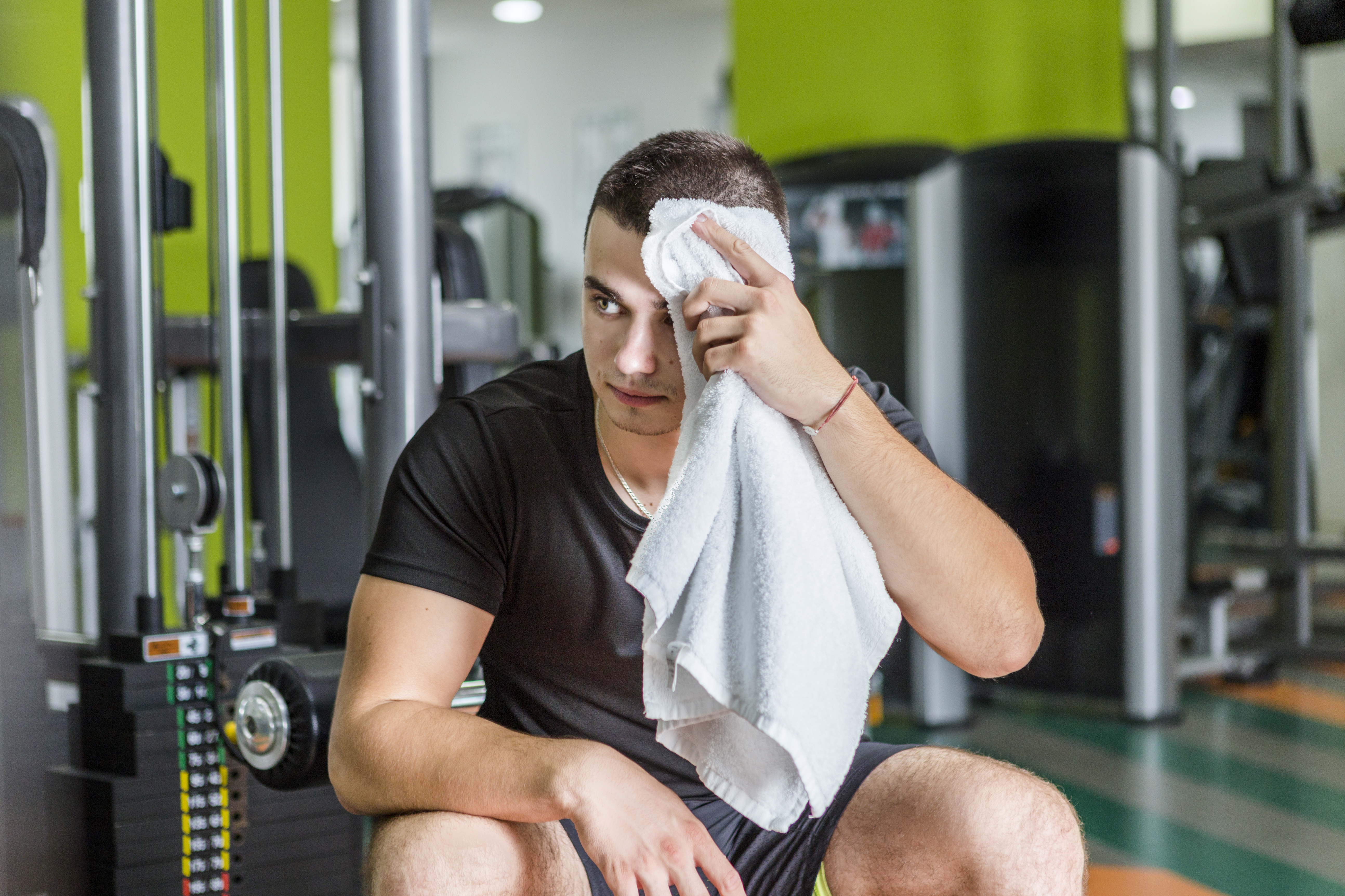 A male athlete sitting in a gym wiping sweat from his face with a white sweat towel for gyms after an intense workout session